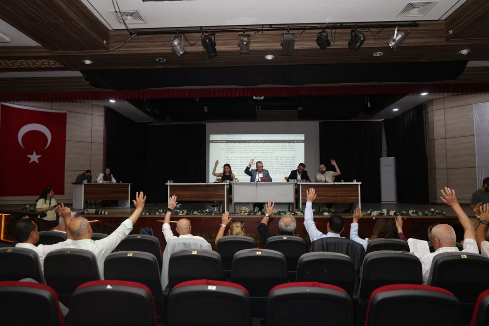 Konferans salonunda düzenlenen bir toplantıda, katılımcıların büyük çoğunluğu oylama yapmak için ellerini havaya kaldırmış durumda. Sahnedeki masada oturan dört kişilik heyet ve salondaki izleyiciler, arkadaki dev ekrana yansıtılan resmi bir metni takip ediyor. Salonun sol tarafında asılı büyük bir Türk bayrağı bulunuyor.