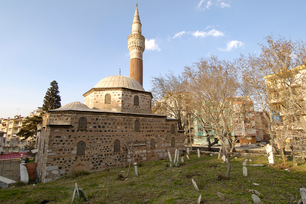 Taş ve tuğla örgülü duvarlara, kurşun kaplı kubbelere ve ince uzun bir minareye sahip tarihi bir cami. Caminin çevresinde, yaprakları dökülmüş ağaçların bulunduğu, eski mezar taşlarının yer aldığı eğimli bir bahçe alanı ve arka planda modern apartmanlar görülüyor.