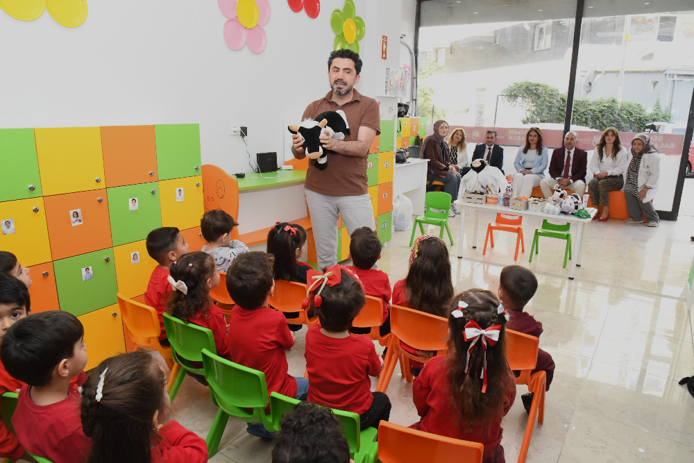 Kahverengi tişörtlü bir eğitmen, anaokulu sınıfında kırmızı tişörtler giymiş çocuklara elindeki inek figürlü oyuncakla bir şeyler anlatıyor. 