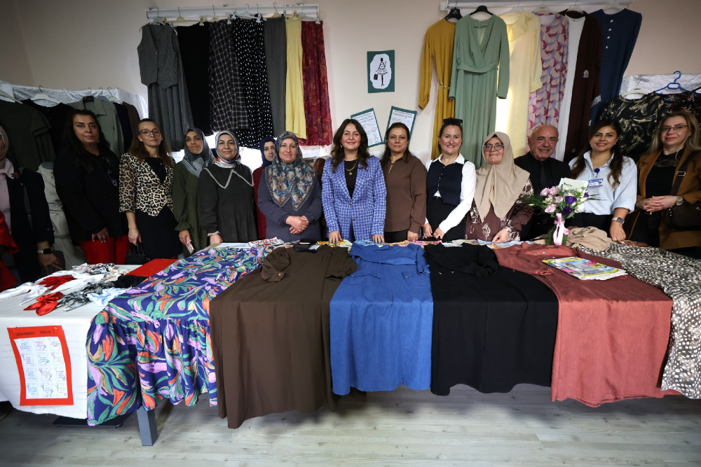 Bir atölye veya sergi alanında, masanın arkasında yan yana dizilmiş on dört kadın ve bir erkekten oluşan grup, önlerindeki masanın üzerine serilmiş çeşitli renk ve desenlerdeki elbiselerle birlikte poz veriyor. 