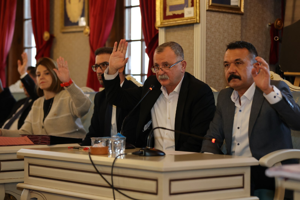 Resmi bir toplantı masasında yan yana oturan dört kişi, oylama yapmak için ellerini havaya kaldırmış durumda.
