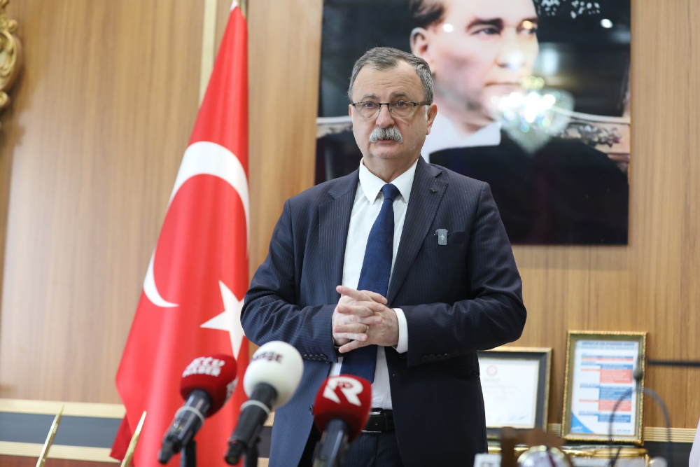 Yunusemre Belediye Başkanı Semih Balaban, lacivert takım elbisesi ve kravatıyla, önünde basın mikrofonlarının bulunduğu bir kürsüde konuşma yapıyor. Arka planda Türk bayrağı ve Mustafa Kemal Atatürk'ün portresi yer alıyor.