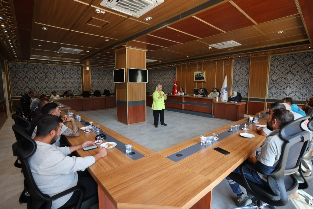 Modern bir toplantı salonunda, açık yeşil ceketli bir kadın ayakta konuşma yaparken, U şeklinde düzenlenmiş masalarda oturan bir grup katılımcı onu dinliyor. 