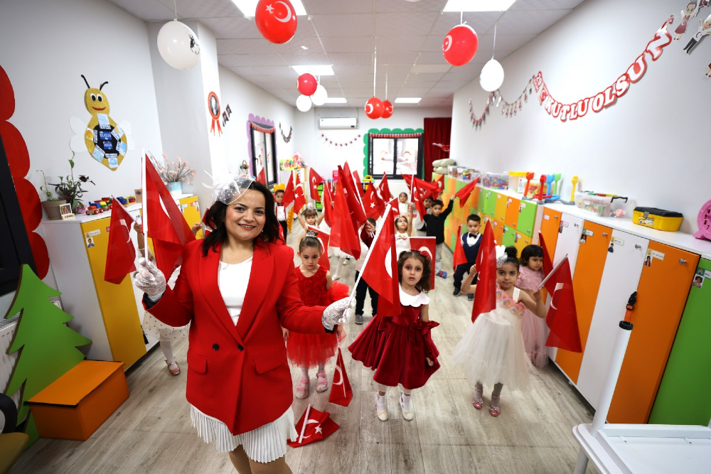 Kırmızı ceketli ve beyaz etekli bir kadın ile ellerinde Türk bayrakları tutan anaokulu öğrencileri, 23 Nisan Ulusal Egemenlik ve Çocuk Bayramı kutlaması için süslenmiş sınıfta bir arada duruyor. Tavanda kırmızı ve beyaz balonların asılı olduğu sınıfta, duvarda 23 Nisan kutlama yazısı ve çocukların yaptığı süslemeler yer alıyor. Öğretmen ve çocuklar neşeyle bayraklarını sallarken, sınıfın duvarlarında renkli dolaplar ve eğitici materyaller bulunuyor.