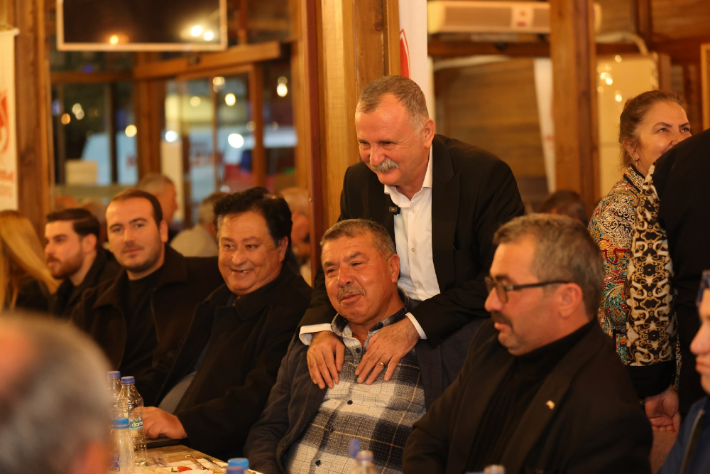 Kapalı bir mekanda, bir masanın etrafında toplanmış bir grup insan yer alıyor. 