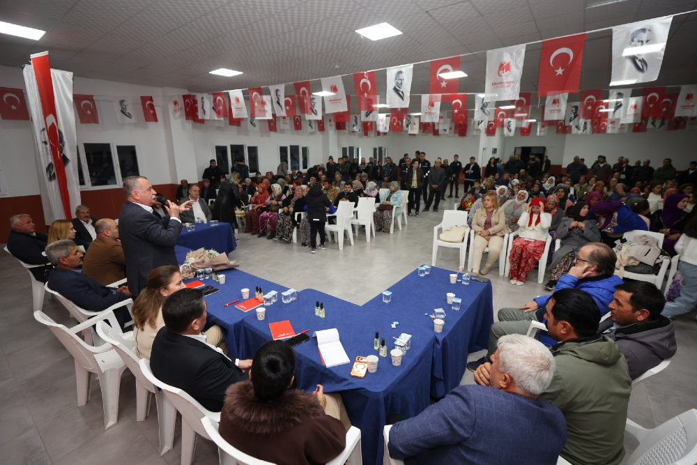Kapalı bir salonda düzenlenen toplantıda, takım elbiseli bir erkek ayakta mikrofonla konuşma yapıyor. 