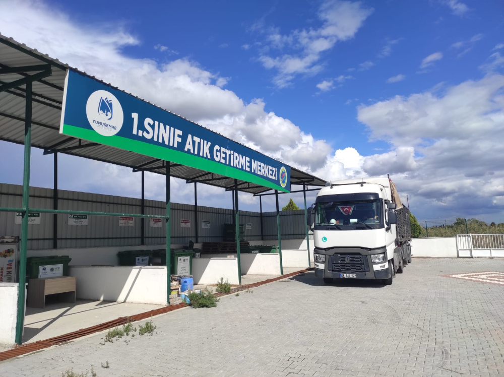 Yunus Emre Belediyesi'ne ait, üzerinde "1. Sınıf Atık Getirme Merkezi" yazan büyük bir tabelanın bulunduğu üstü kapalı alanın önünde park halinde duran beyaz bir kamyon. Açık havada, kilitli parke taşlı bir zemin üzerinde yer alan merkezin yan tarafında atık toplama konteynerleri ve çeşitli atık bölmeleri bulunuyor.