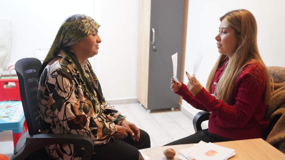Başörtülü orta yaşlı bir kadın ile karşısında oturan uzun sarı saçlı ve kırmızı kazaklı genç bir kadın, bir masa etrafında oturuyor. 