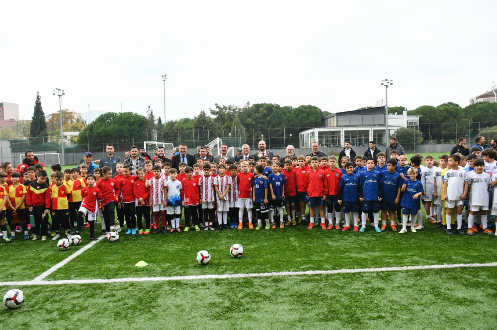 Çeşitli futbol takımı formaları giymiş çok sayıda çocuk ve arkalarında duran yetişkinlerden oluşan geniş bir grup, yeşil bir futbol sahasında yan yana dizilmiş poz veriyor. 