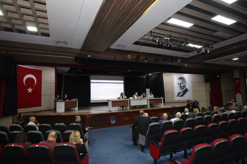 Modern bir konferans salonunda düzenlenen panelde, sahnedeki uzun bir masada oturan beş konuşmacı ve yan taraftaki küçük bir masada iki görevli yer alıyor. 