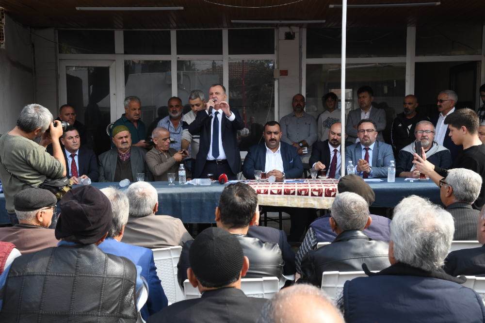 Açık havada, bir masa etrafında toplanmış kalabalık bir gruba hitap eden, takım elbiseli ve kravatlı bir adam mikrofonla konuşma yapıyor.